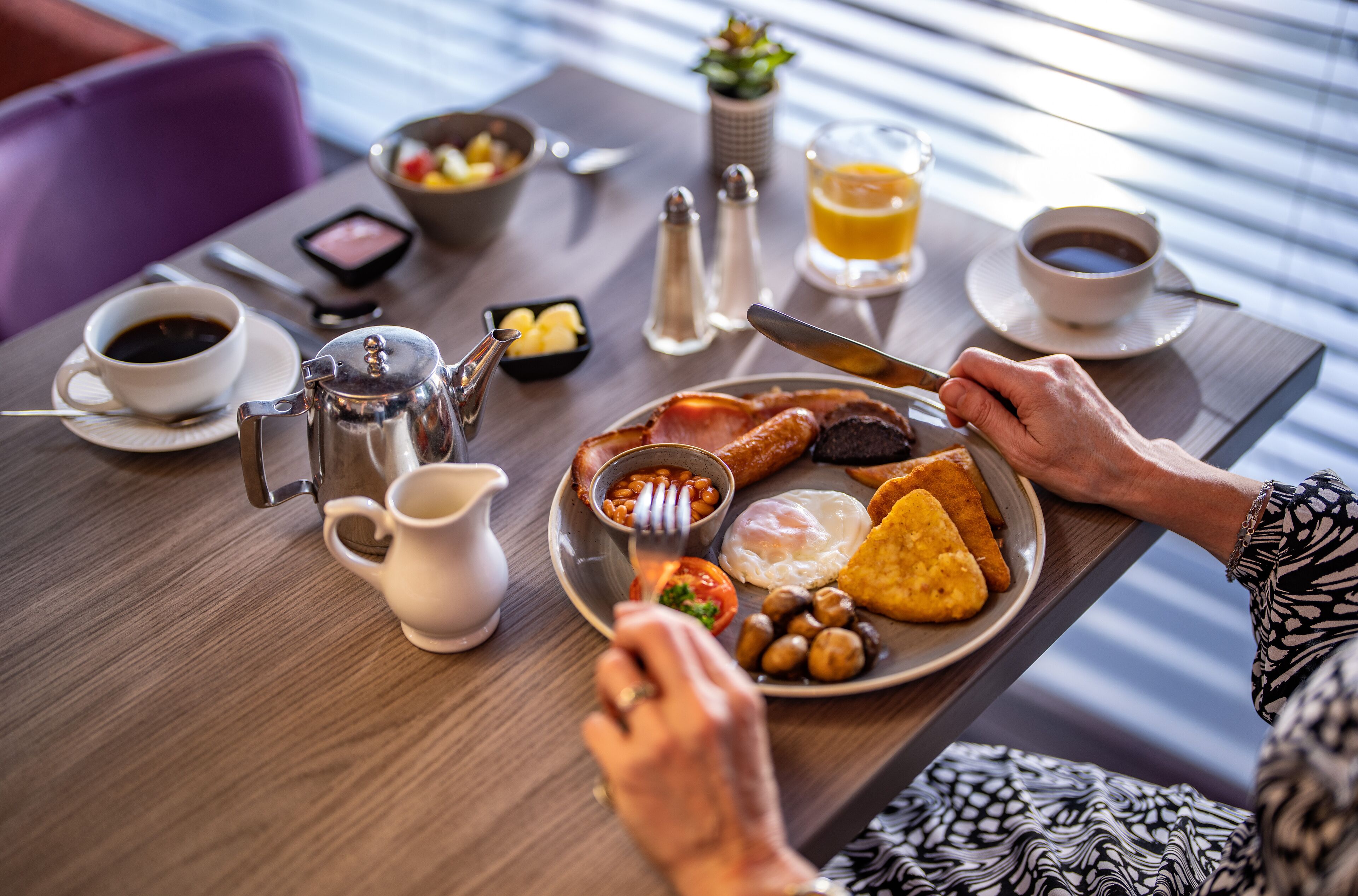 Une table dressée pour le petit-déjeuner avec une assiette contenant des œufs, du bacon, des champignons et des toasts, une tasse de café, un verre de jus d’orange et un pichet de lait, le tout posé sur une table en bois.