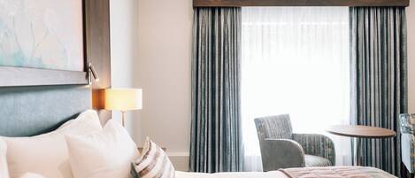 A hotel room with a bed, a chair, a small table, and a painting on the wall.