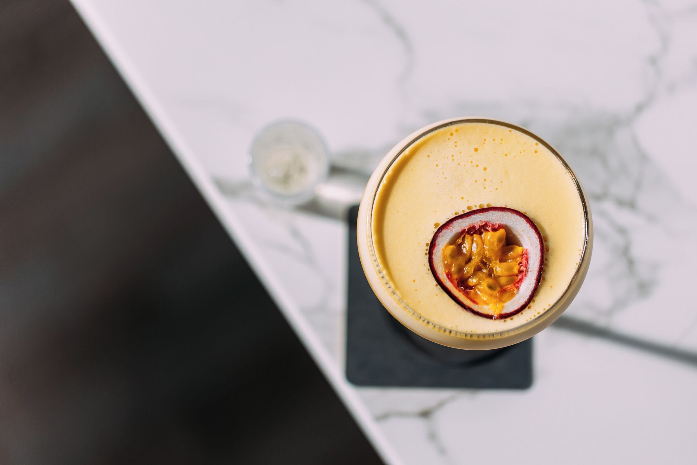 Vista cenital de una mesa con un vaso que contiene una bebida amarilla y una rodaja de maracuyá.