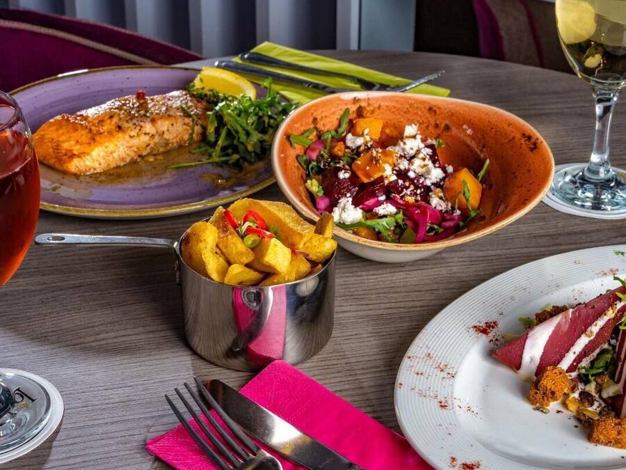 Uma mesa de jantar com diversos pratos, incluindo um prato de peixe, uma salada numa tigela e um pequeno recipiente de metal com batatas fritas.