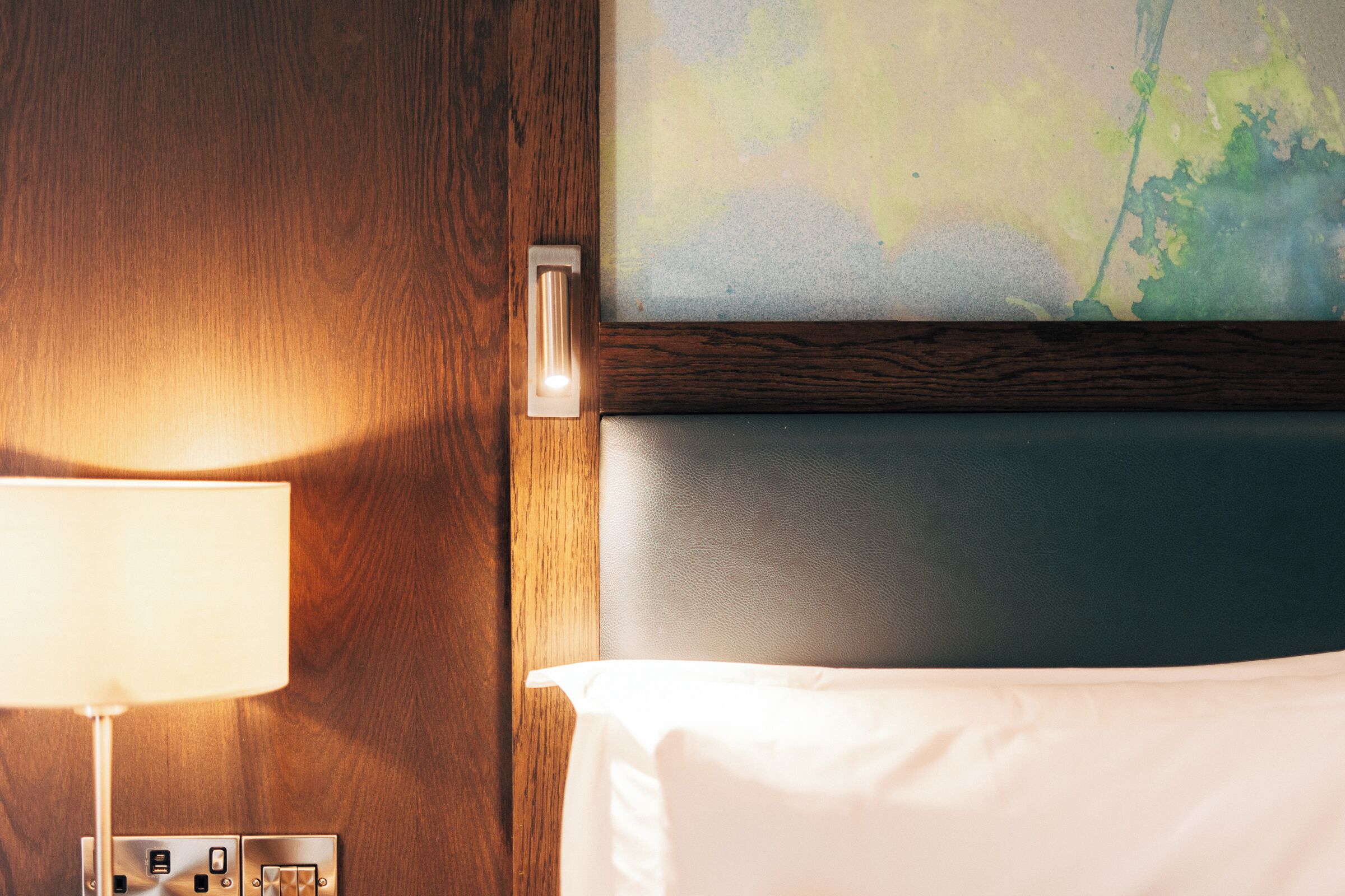 A hotel room with a wooden wall, a white bed, a lamp, and a painting above the bed.