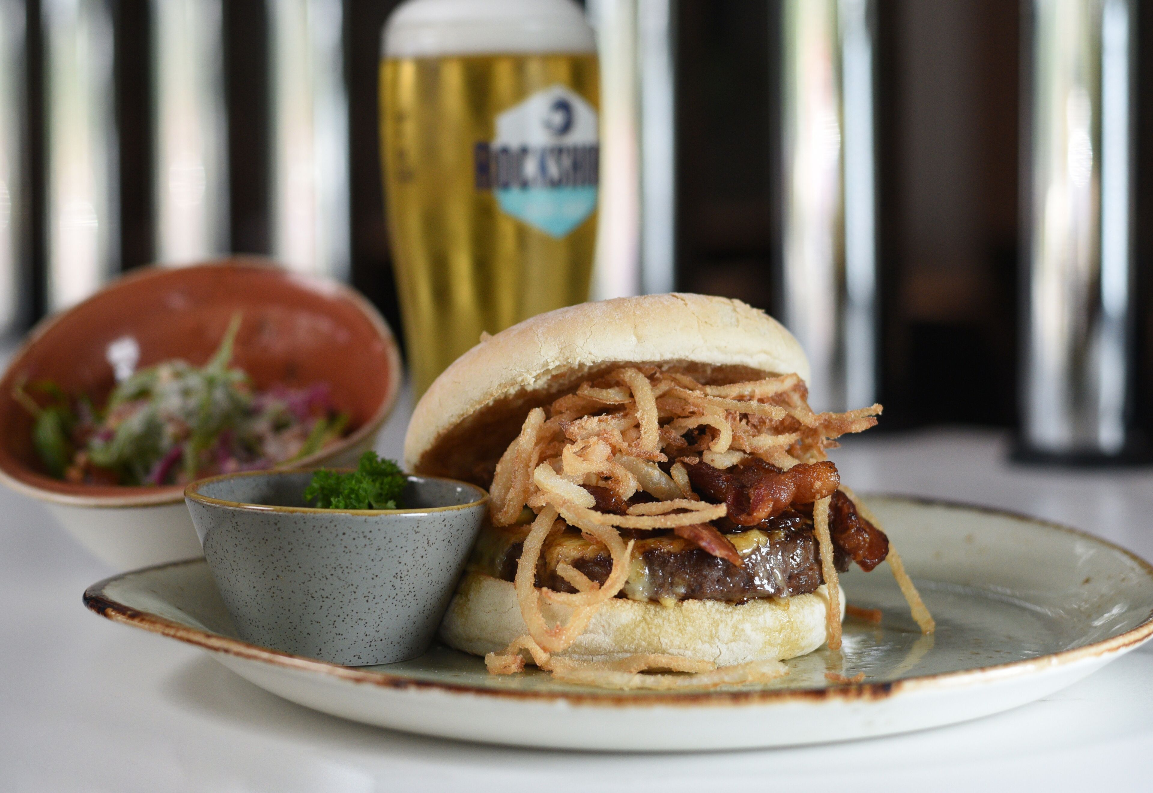 Una hamburguesa con tocino crujiente y cebollas, un cuenco pequeño de ensalada y una jarra de cerveza sobre un plato.