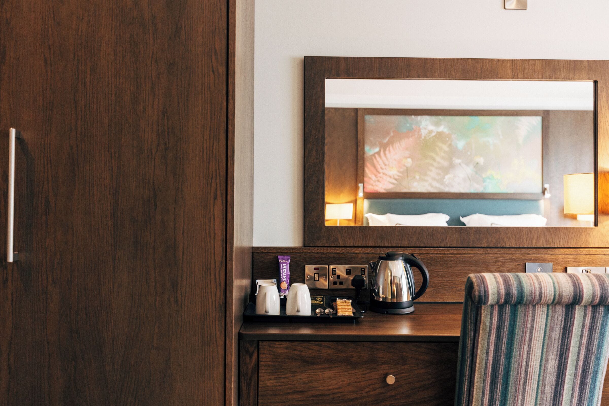 A hotel room with a wooden wardrobe, a desk with a coffee maker, a striped chair, and a mirror reflecting a painting.