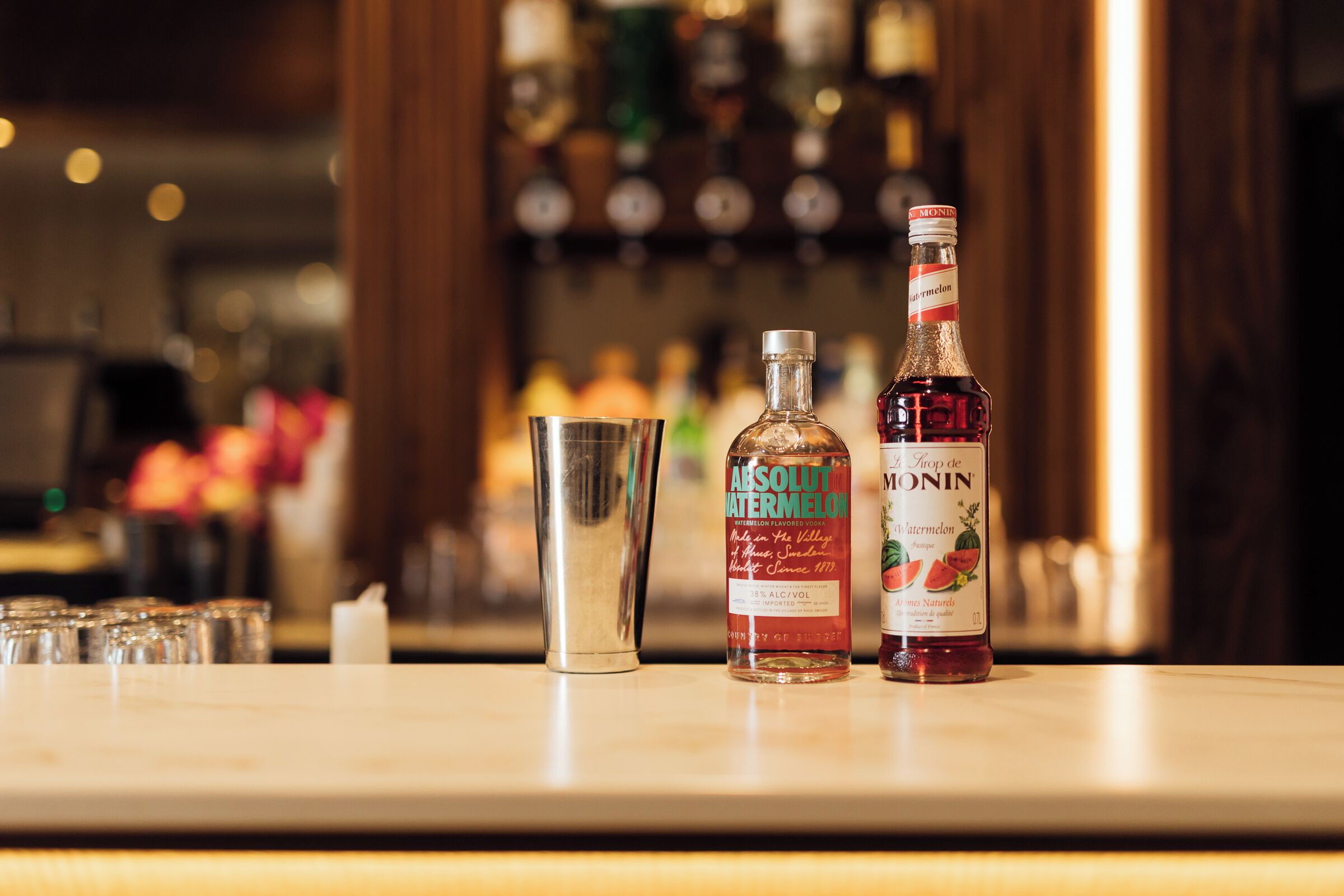 A bar counter with two bottles of Absolut watermelon and Monin, a cocktail shaker, and several glasses.