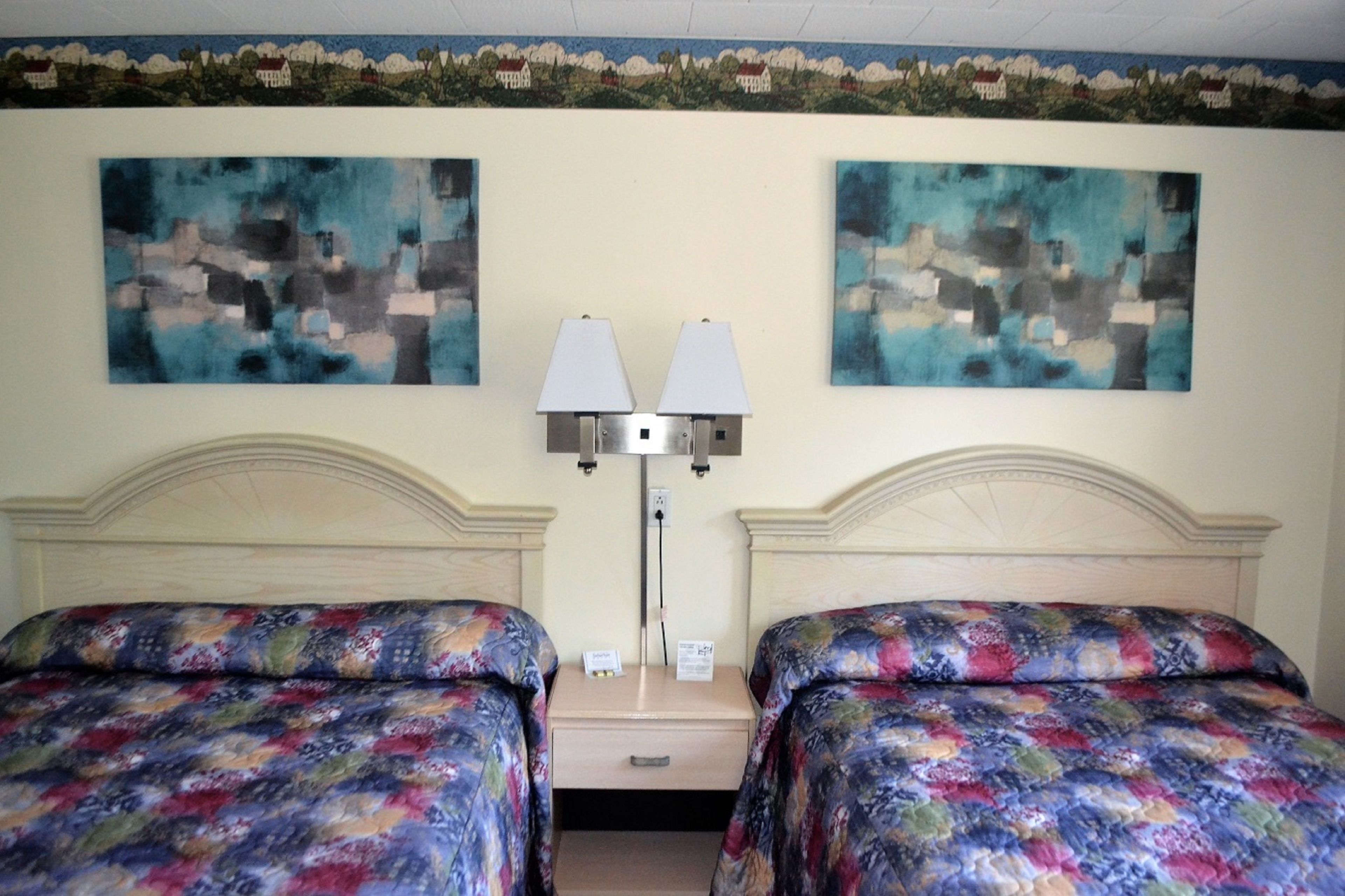 A hotel room with two beds, each with a floral-patterned bedspread, a nightstand with a lamp, and two abstract paintings on the wall.