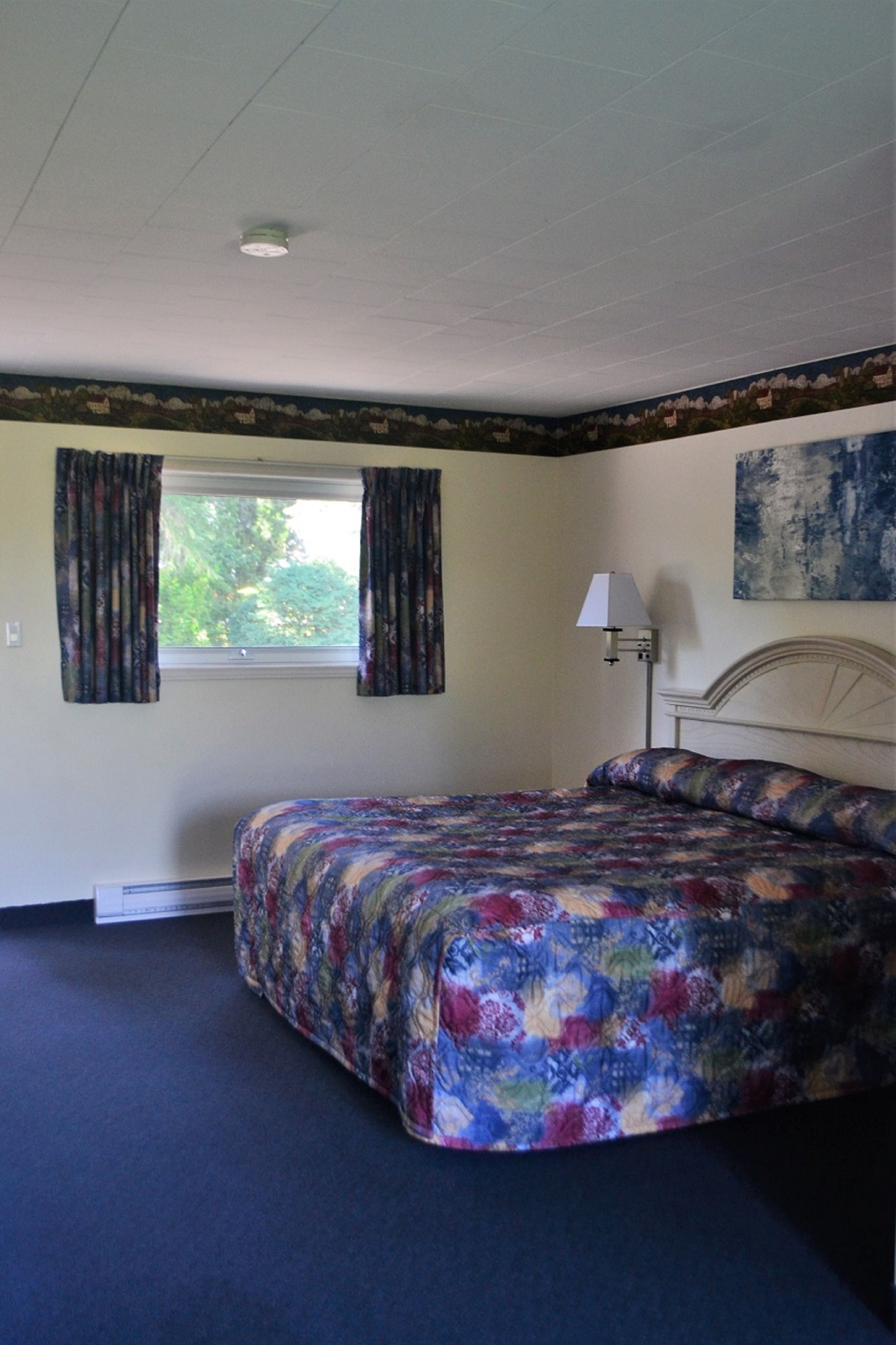 Um quarto com cama, janela com cortinas, abajur e um quadro na parede.