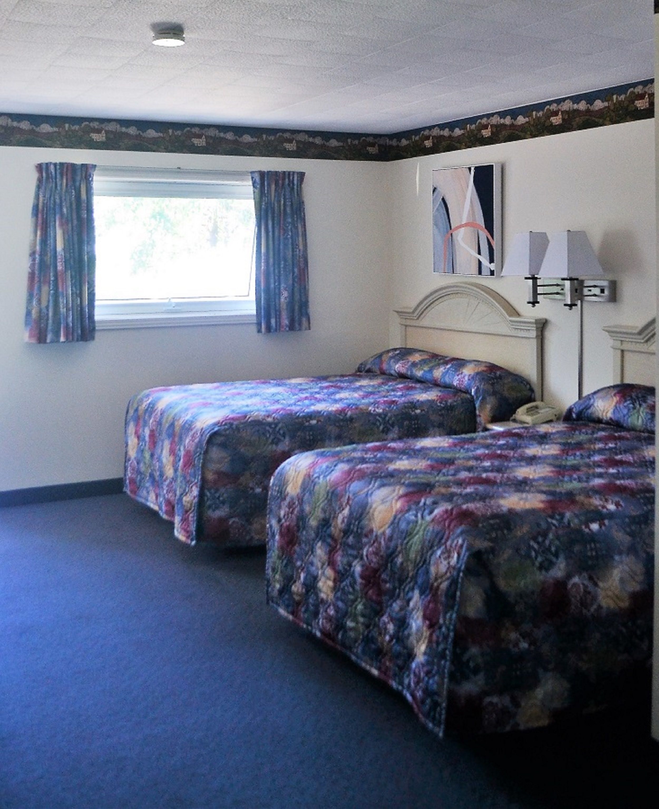 A room with two beds, a window with curtains, a lamp, and a wall with a framed picture.