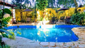 Una piscina con sillas de descanso y sombrillas, rodeada de follaje y un muro amarillo.