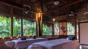 A room with two beds, large windows with a view of greenery, a thatched roof, and a decorative wall hanging.