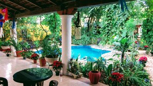 A covered outdoor area with a pool, surrounded by various potted plants and a table with chairs.