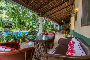 Un área de estar al aire libre con techo y vista a la piscina, amueblada con sillones de cuero y una mesa de vidrio.