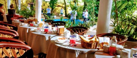 Un comedor con mesas redondas, sillas de mimbre y una piscina al fondo.