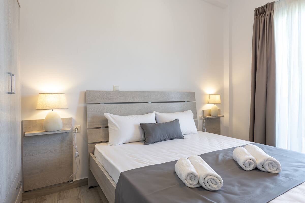 Un lit bien fait, avec du linge de lit blanc et gris, deux lampes de chevet et des serviettes pliées posées sur le lit.