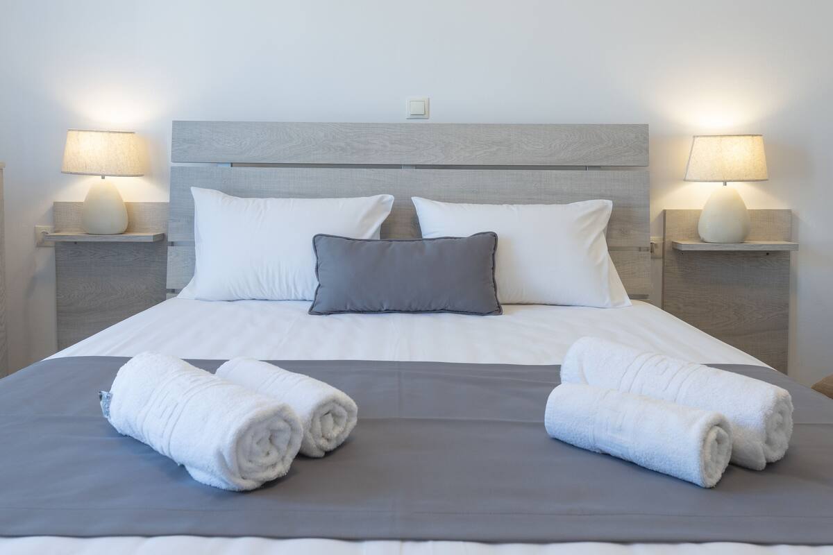 Un lit bien fait, avec du linge de lit blanc et gris, deux lampes de chevet et un interrupteur placé sur la tête de lit.