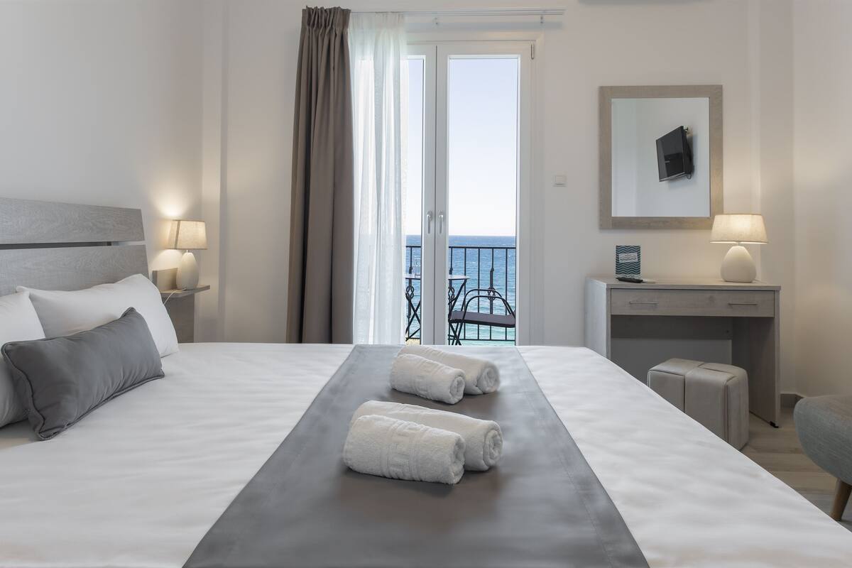Une chambre d’hôtel avec un lit, deux serviettes, un bureau, une chaise, une lampe et une vue sur la mer, visible à travers une porte vitrée.