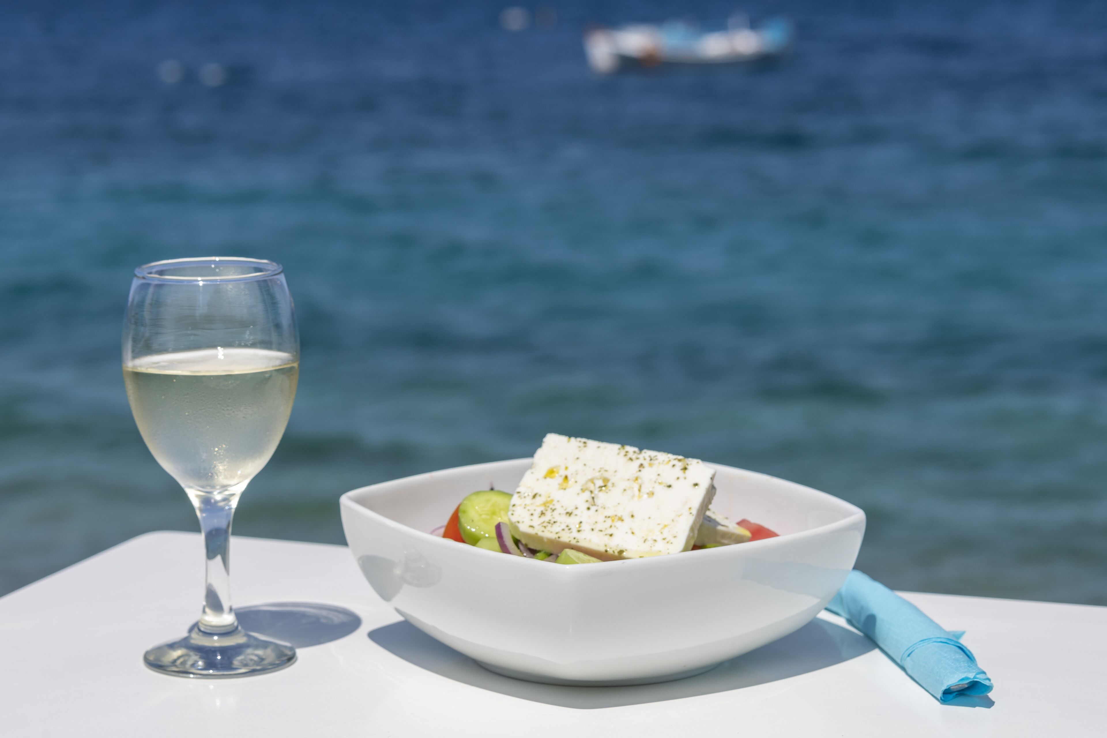 A white table with a white bowl containing a salad and a glass of white wine, set against a backdrop of a blue sea and a boat.