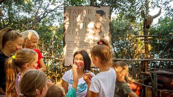 A group of children gathered around a sign in a natural setting with trees and a wooden structure in the background.