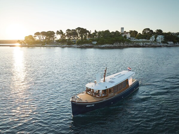 A boat with the number 895 PO on its side, sailing on calm waters with a clear sky and a distant shoreline with trees and buildings. -  Isabella Island Miramare Suites - Villas, Valamar Collection (Porec)