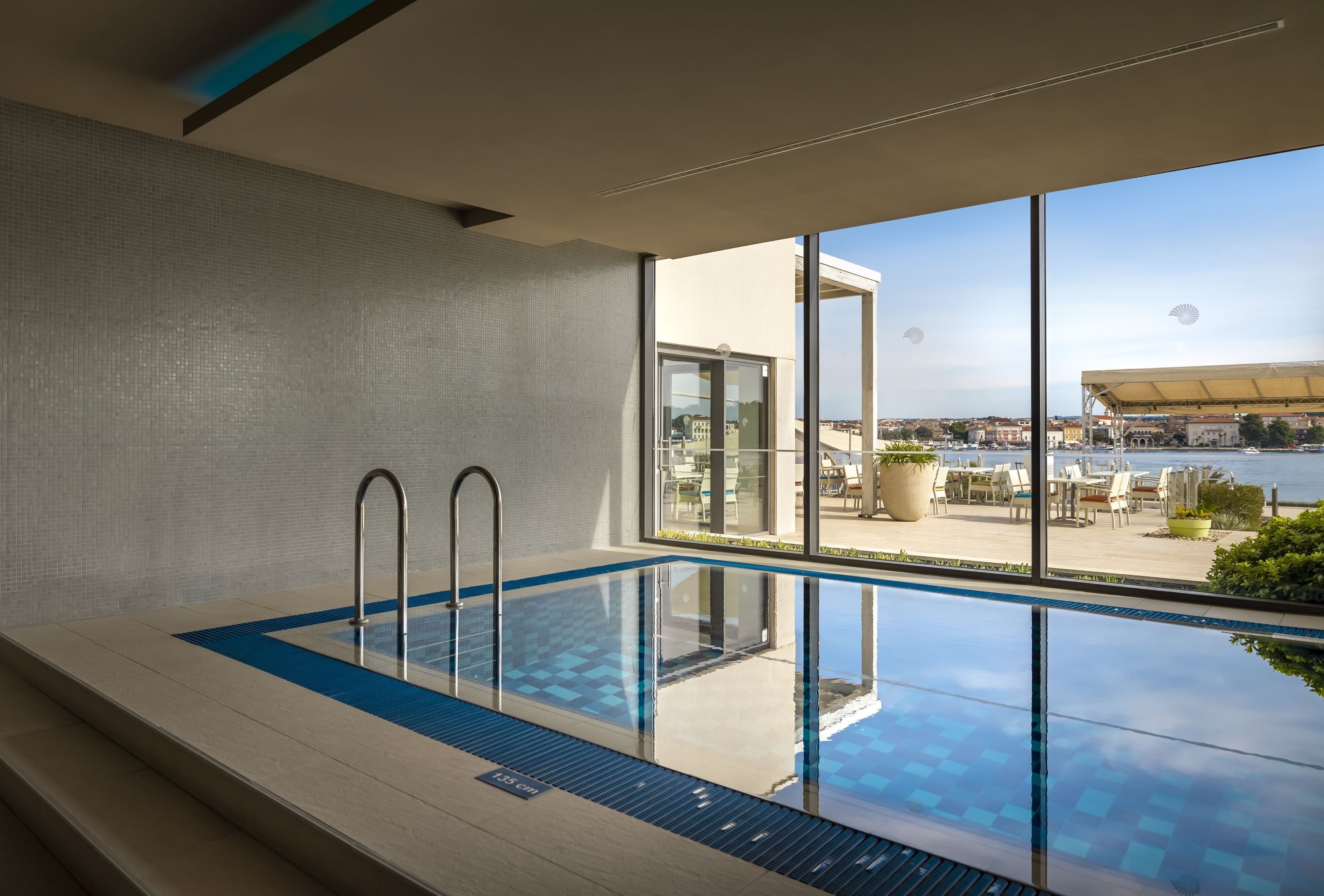 An indoor swimming pool with clear glass walls, a view of a cityscape, and a patio area with outdoor seating.