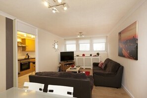 Un espacio habitable compacto con comedor, sofá, televisión y cocina al fondo.