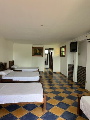 Habitación de hotel con cuatro camas individuales, piso de cuadros, un cuadro en la pared y un televisor montado.