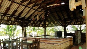 Un espacio exterior cubierto con mesas y sillas de madera, una barra de ladrillo y vistas a palmeras.