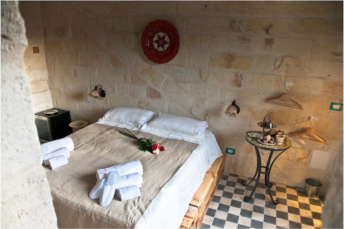 A bedroom with a checkered floor, a bed with white linens, a small table with a tray of food, and a red decorative plate on the wall.