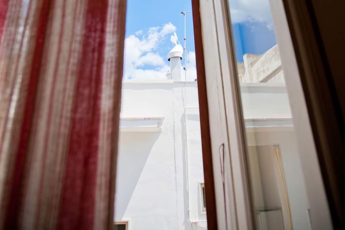 A view through a window with a striped curtain, revealing a building with a white facade and a clear blue sky with clouds.