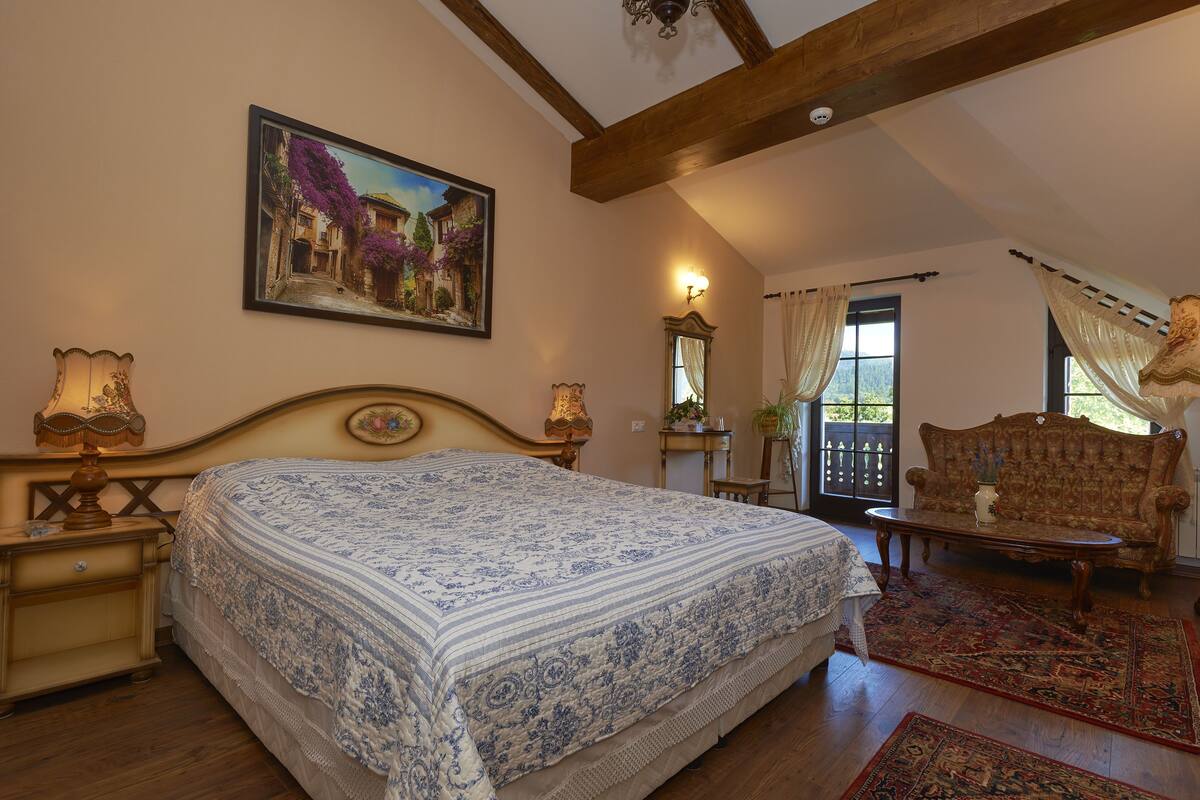 Un dormitorio con una cama grande, cabecera de madera, un cuadro en la pared, una mesita de noche con lámpara, un aparador, un sofá y una ventana con cortinas.