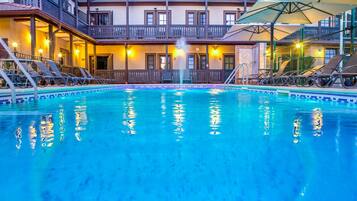 A swimming pool with lounge chairs and umbrellas, surrounded by a building with balconies and lights.