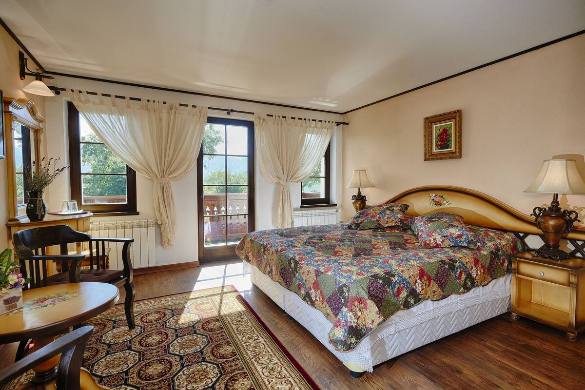 Un dormitorio con una cama grande, una mesita de noche de madera, una mesa con sillas y una ventana con cortinas.