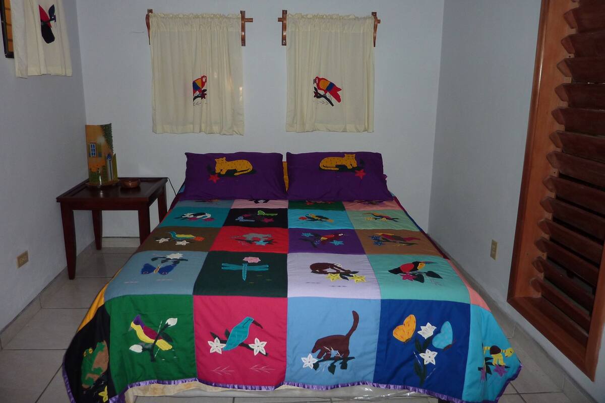 Um quarto com um edredom colorido, duas almofadas roxas, uma pequena mesa de cabeceira e uma janela com cortinas.