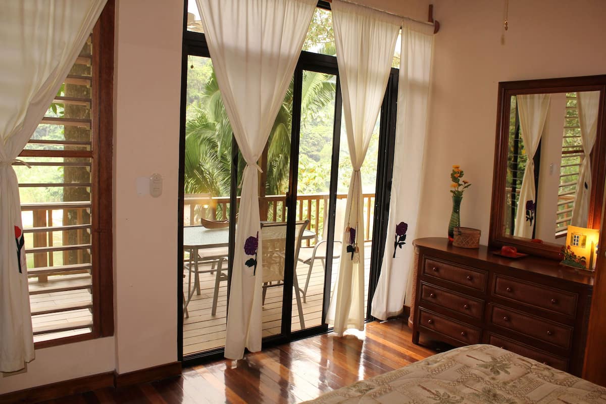 Quarto com piso de madeira, cama com colcha florida, cômoda e espelho. Há uma porta de vidro deslizante que dá para uma área externa com cadeiras e uma mesa.