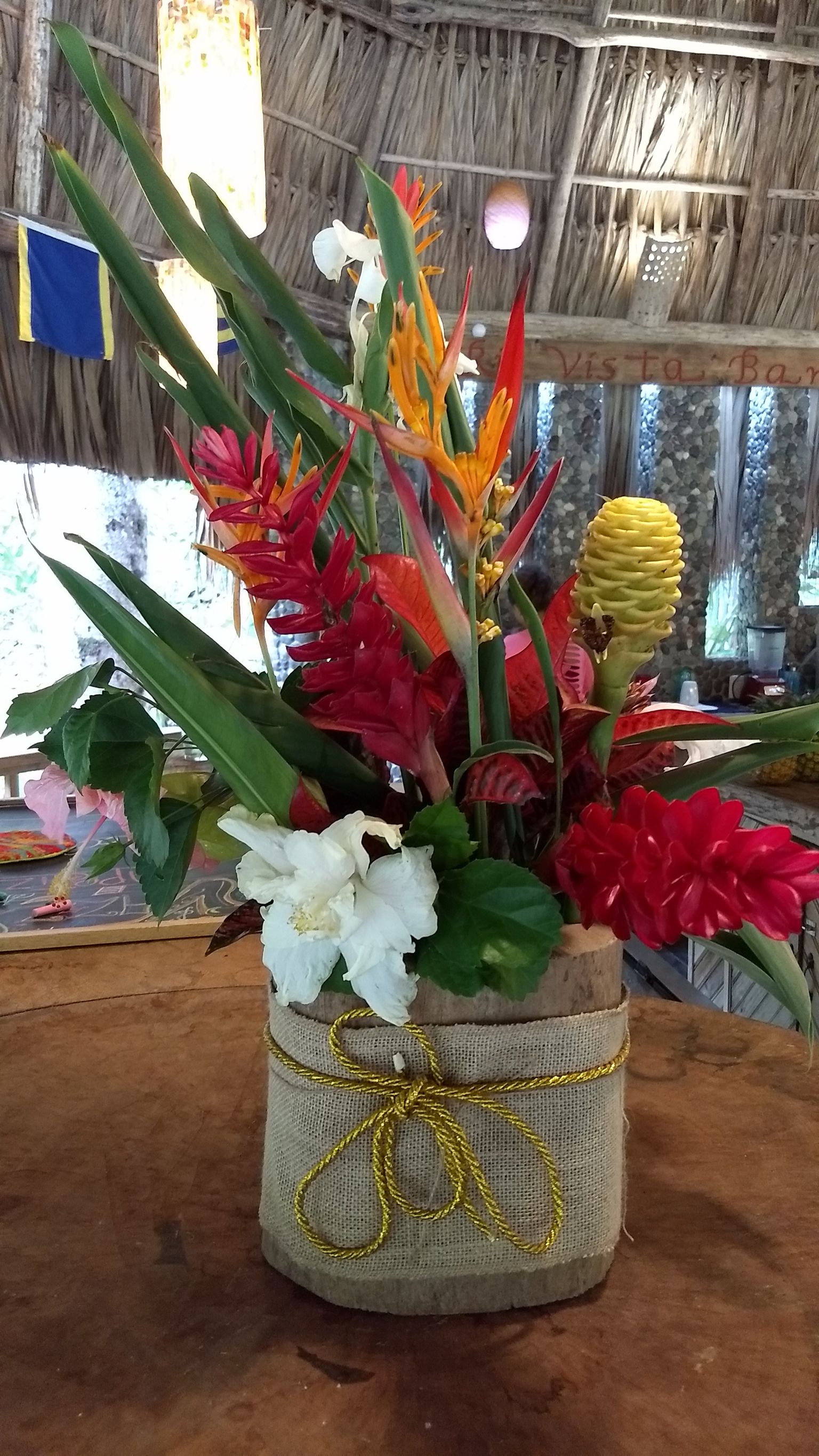 A vase with a burlap base and a golden ribbon, containing a variety of flowers including red, white, and yellow blooms, set against a rustic backdrop.