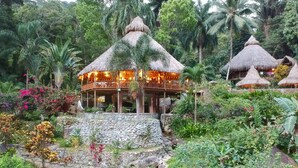 A thatched-roof structure with a balcony, surrounded by lush greenery and a stone pathway. - Villas Pico Bonito (La Ceiba)