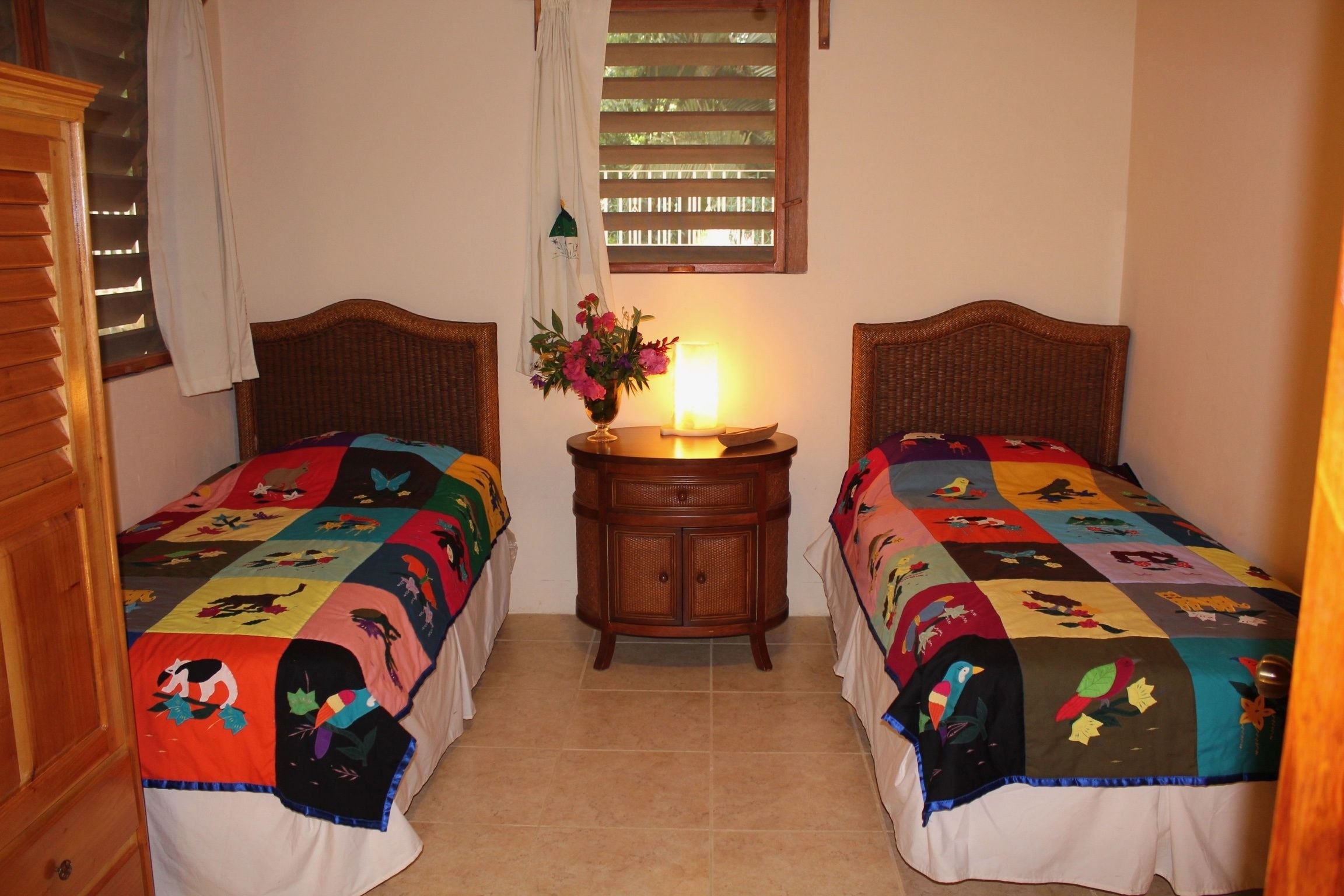 Habitación con dos camas individuales, cada una con un colcha colorida, una mesita de noche con lámpara y una ventana con persianas.