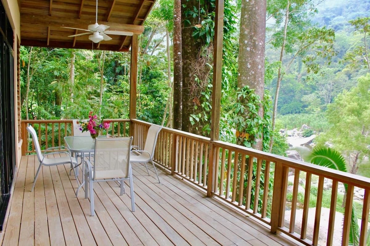 Um deck de madeira com mesa e cadeiras, cercado por um corrimão de madeira, com vista para uma paisagem verde e exuberante.