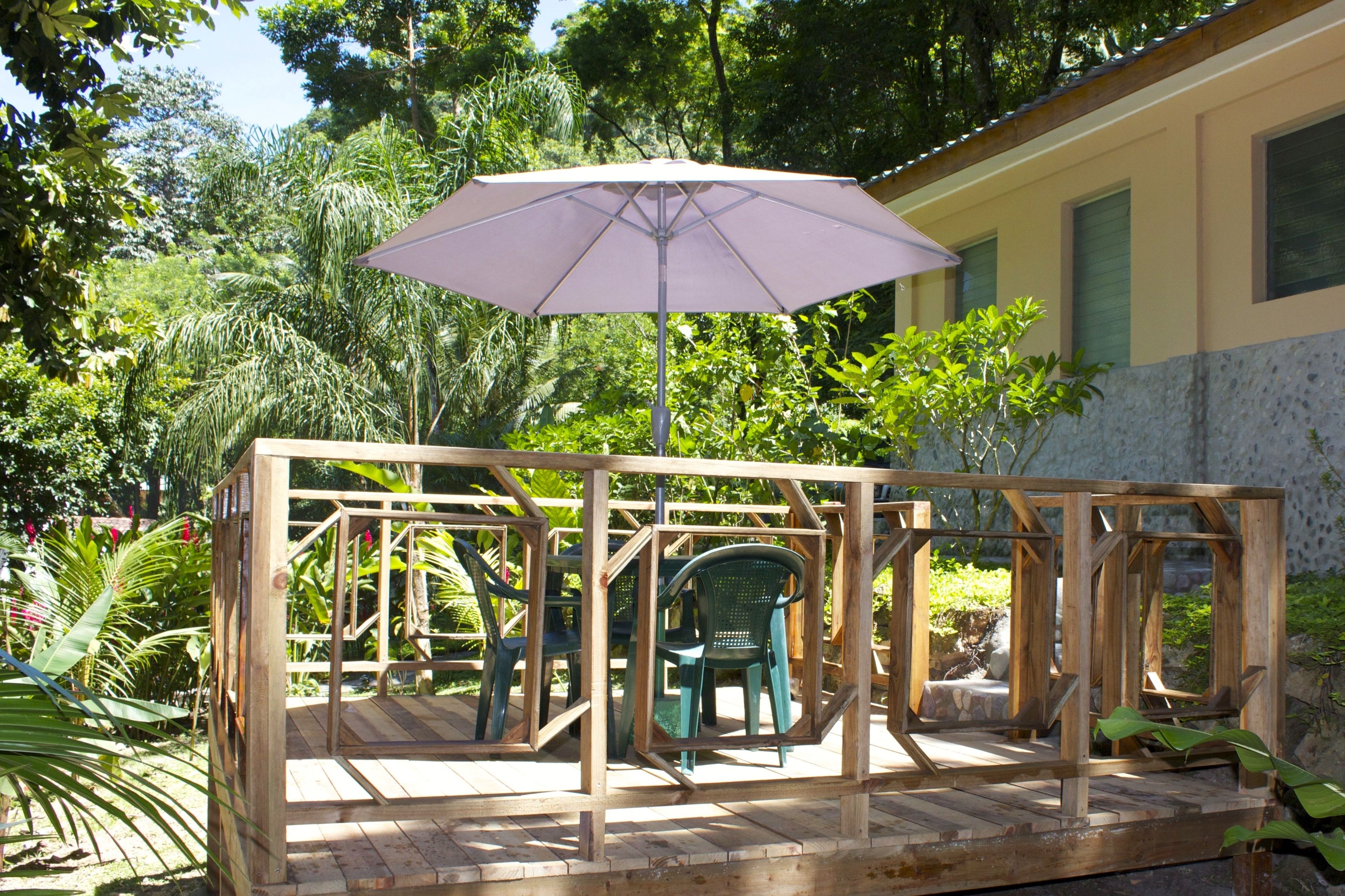 A wooden deck with a purple umbrella, surrounded by greenery and a building in the background.
