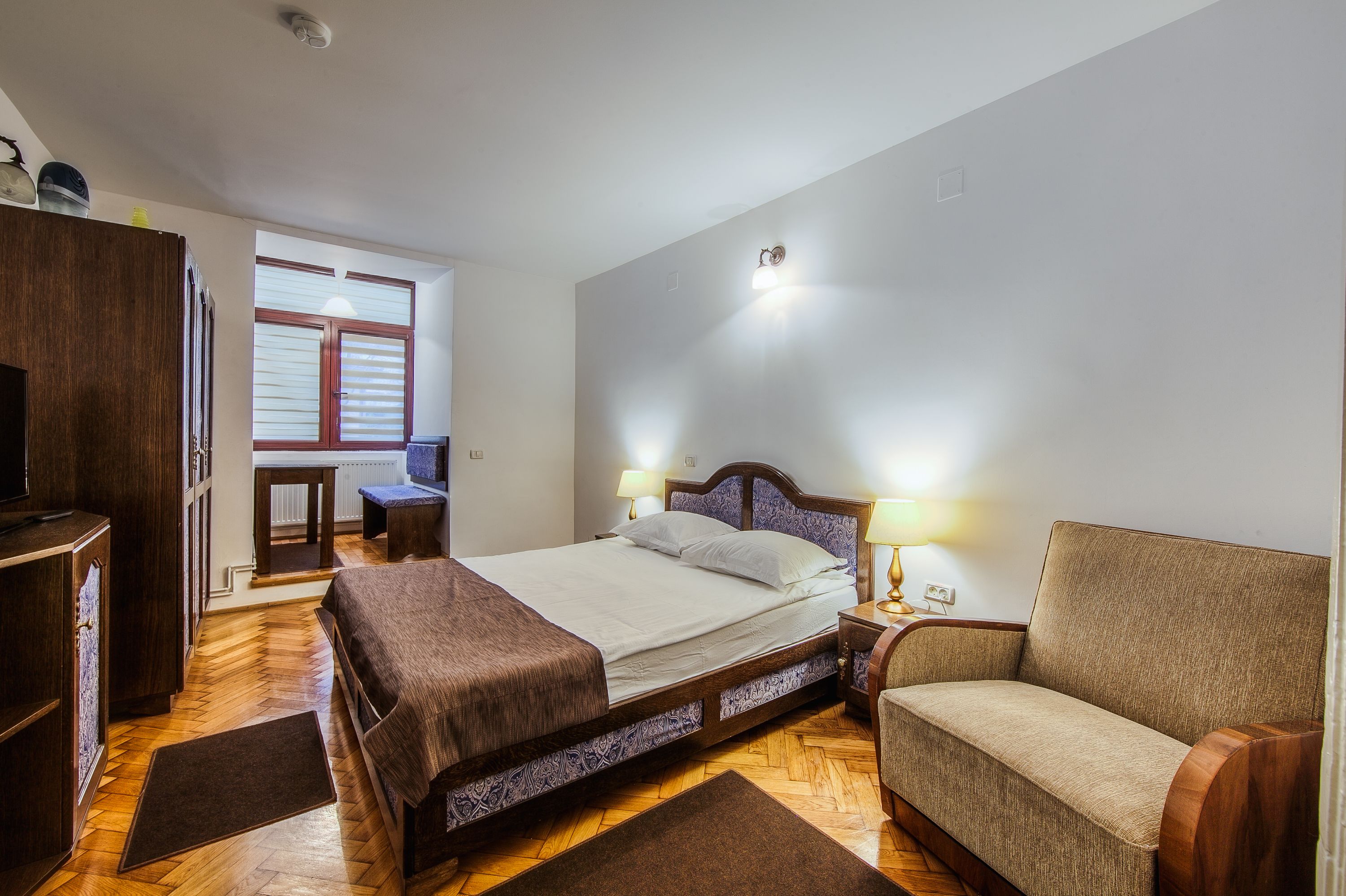 A bedroom with a bed, bedside table, chair, wardrobe, and a window with blinds.