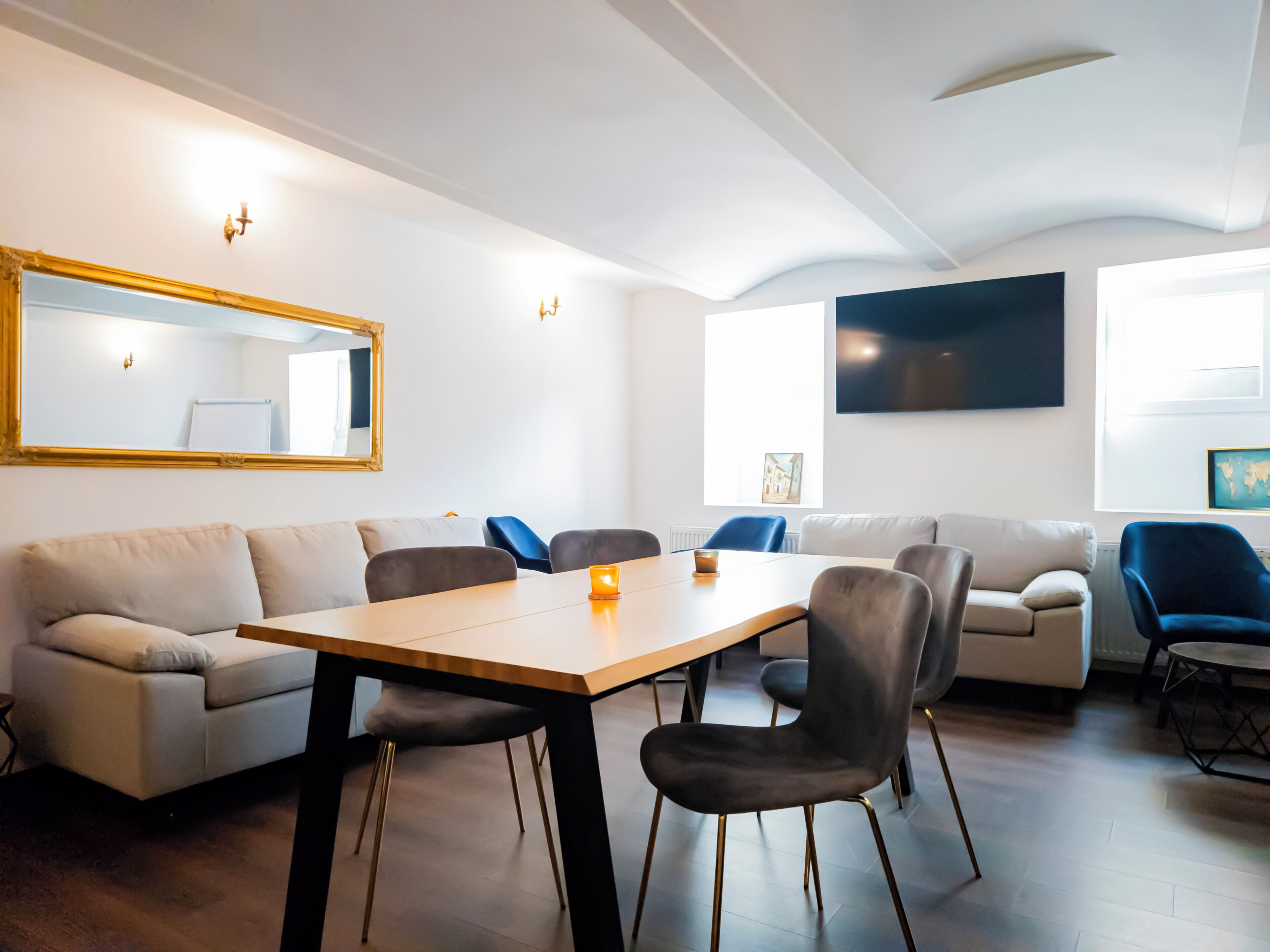 A modern living room with a wooden dining table, grey sofas, and a large mirror.