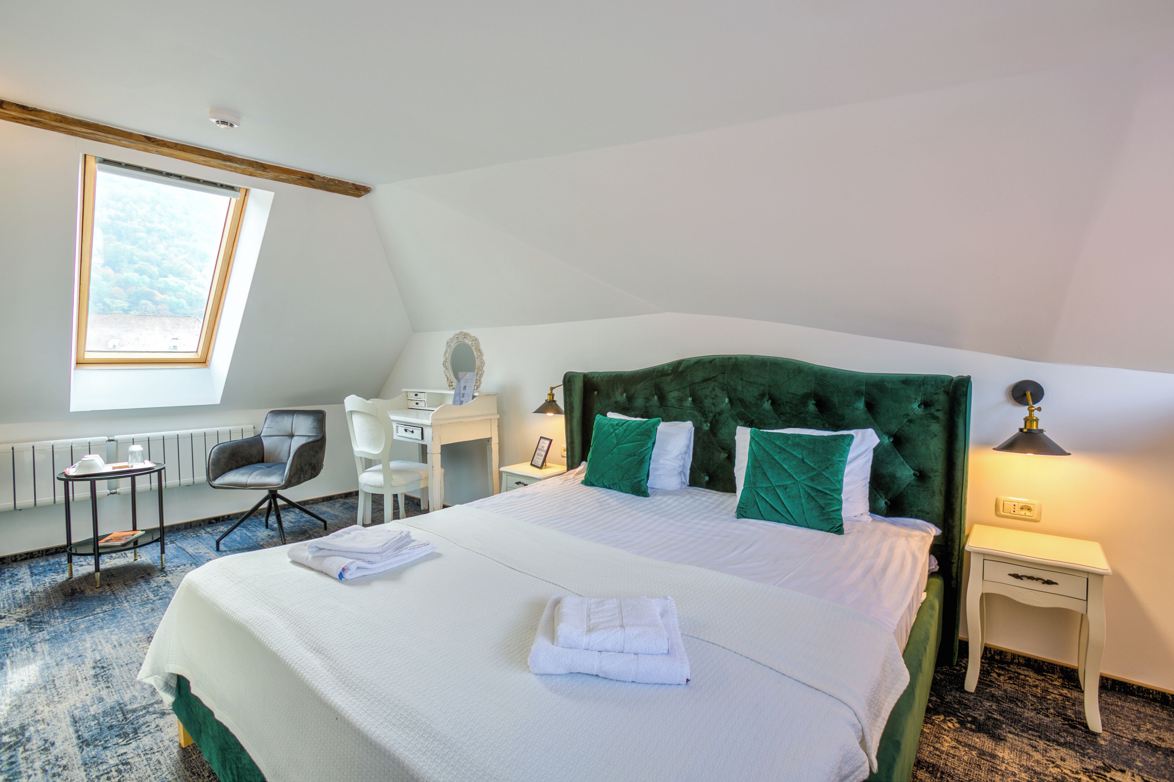 A neatly made bed with green and white bedding, a desk with a chair, a small table with a lamp, and a window with a view of the sky.