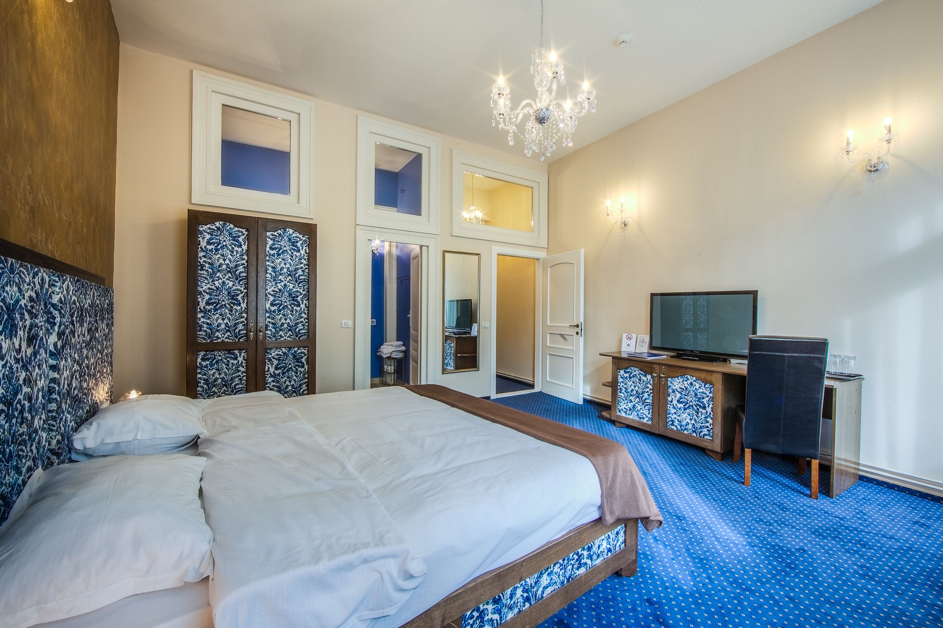 A hotel room with a large bed, a desk with a television, and a chandelier.