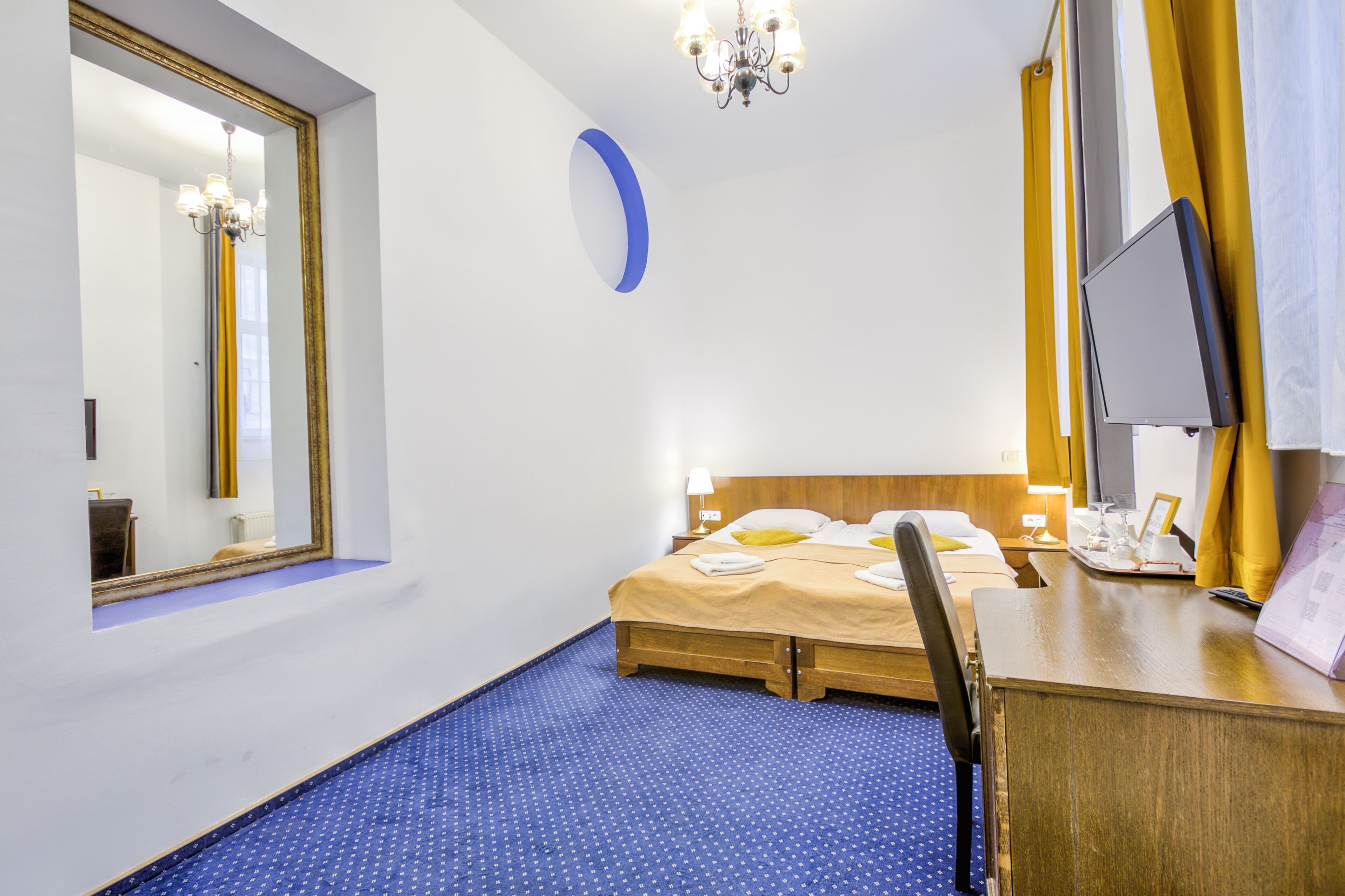 A hotel room with a bed, a desk with a computer, a chair, a mirror, a chandelier, and a blue carpet.