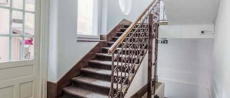 Una escalera con barandilla de madera y alfombra estampada, que conduce a un rellano con una puerta y ventanas.