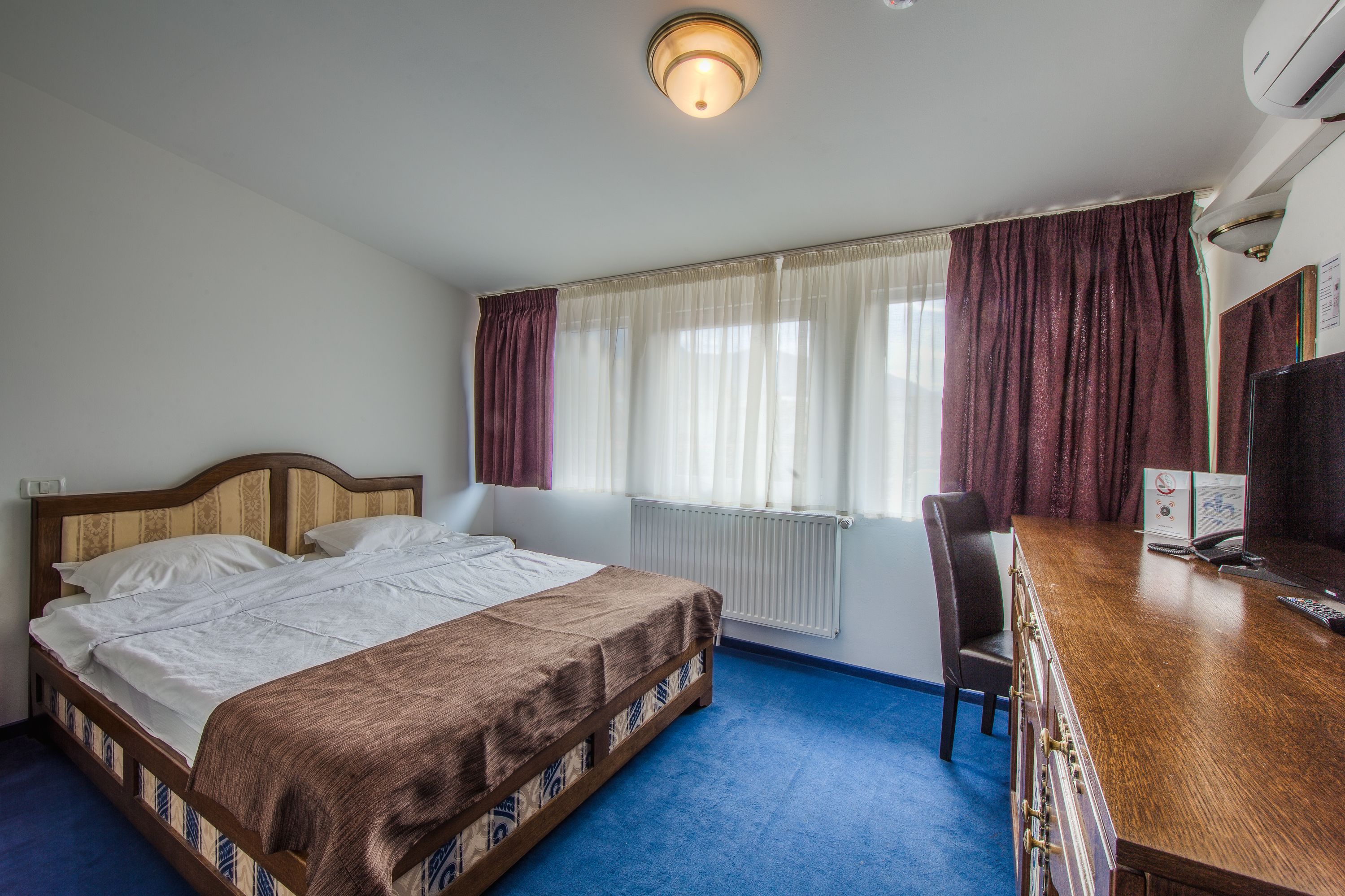 A hotel room with a bed, a desk with a computer, a chair, a window with curtains, and a radiator.