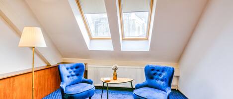 A room with two blue armchairs, a small round table with a vase, a floor lamp, and two skylights.