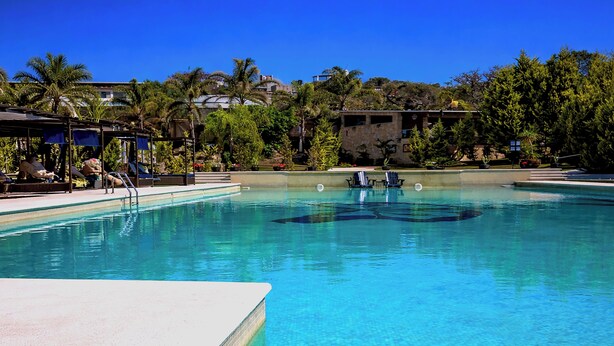 Piscina azul e límpida com espreguiçadeiras e guarda-sóis, cercada por palmeiras e edifícios.