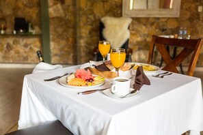 A breakfast table set with a plate of fruits, two glasses of orange juice, a cup of coffee, and a bottle of wine.