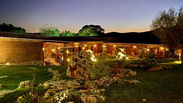 A building with a brick facade, a covered outdoor seating area with string lights, and a landscaped garden with rocks and flowers.