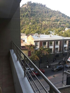 Un balcón con barandilla de metal que da a una calle con coches y edificios, y una colina con árboles al fondo.